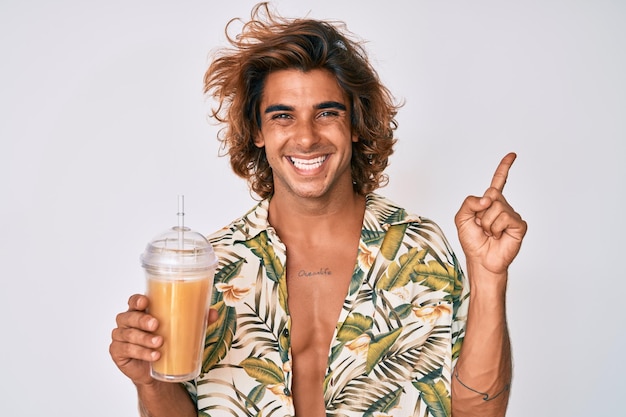 Молодой латиноамериканец в летней рубашке пьет стакан апельсинового сока, счастливо улыбаясь, указывая рукой и пальцем в сторону