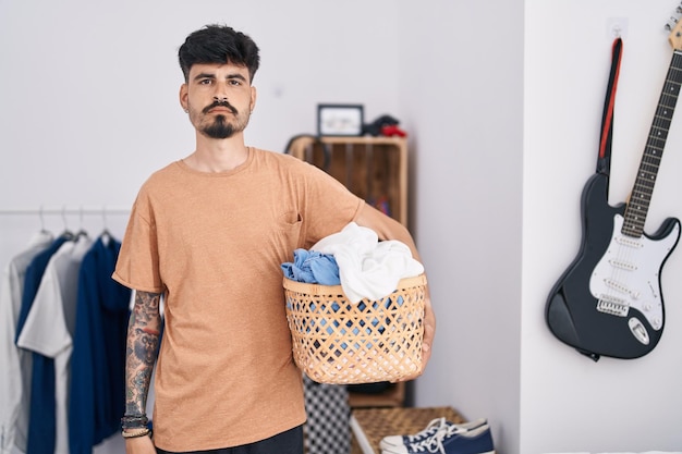 Young hispanic man with beard holding laundry basket at bedroom thinking attitude and sober expression looking self confident