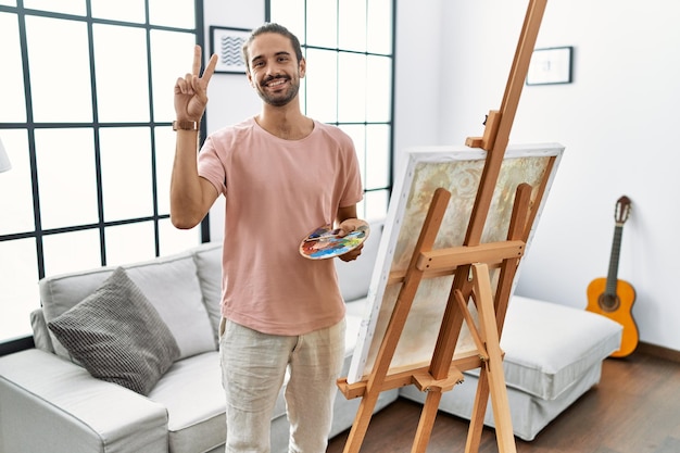 Photo young hispanic man with beard painting on canvas at home smiling with happy face winking at the camera doing victory sign with fingers. number two.