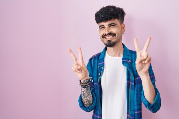 Молодой латиноамериканец с бородой стоит на розовом фоне и улыбается, глядя в камеру, показывая пальцами знак победы номер два