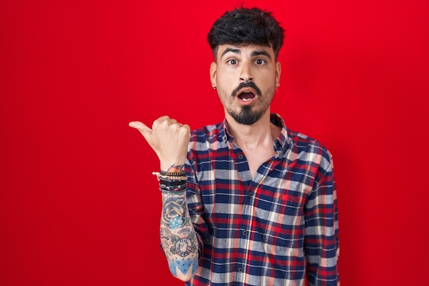 Young hispanic man with beard standing over red background surprised pointing with hand finger to the side open mouth amazed expression