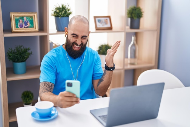 Giovane ispanico con barba e tatuaggi che fa una videochiamata con lo smartphone urlando orgoglioso di celebrare la vittoria e il successo molto eccitato con il braccio alzato