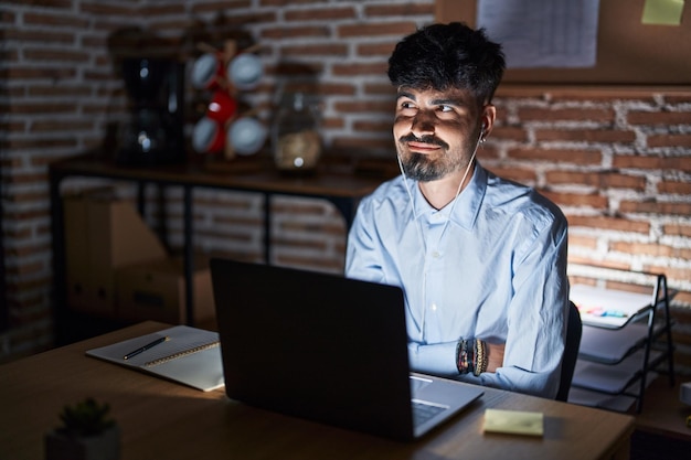 Young hispanic man with beard working at the office at night smiling looking to the side and staring away thinking