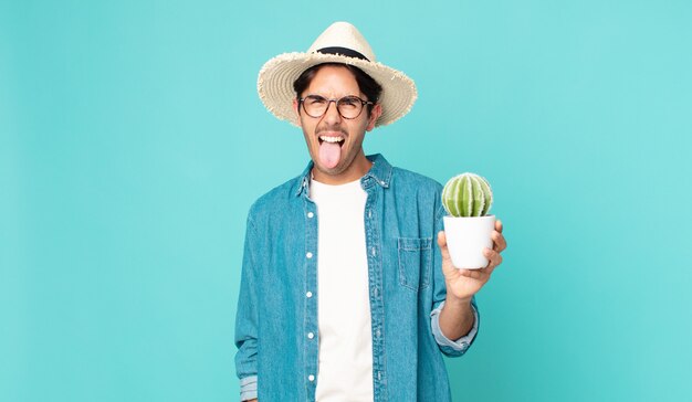 Young hispanic man with cheerful and rebellious attitude, joking and sticking tongue out and holding a cactus