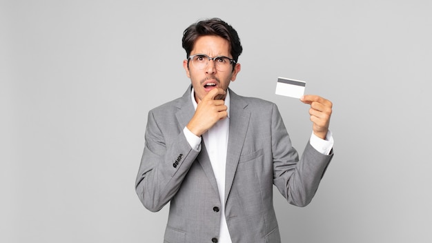 Young hispanic man with mouth and eyes wide open and hand on chin and holding a credit card