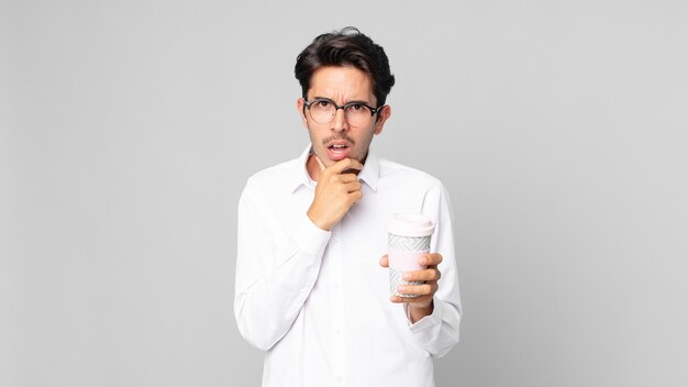 Young hispanic man with mouth and eyes wide open and hand on chin and holding a take away coffee