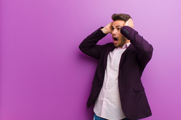 Young hispanic man with open mouth, looking horrified and shocked because of a terrible mistake, raising hands to head