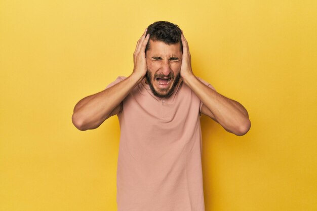 黄色い背景のヒスパニック系の若い男性が ⁇ あまり大きな音を聞かないように手で耳を覆っています ⁇ 
