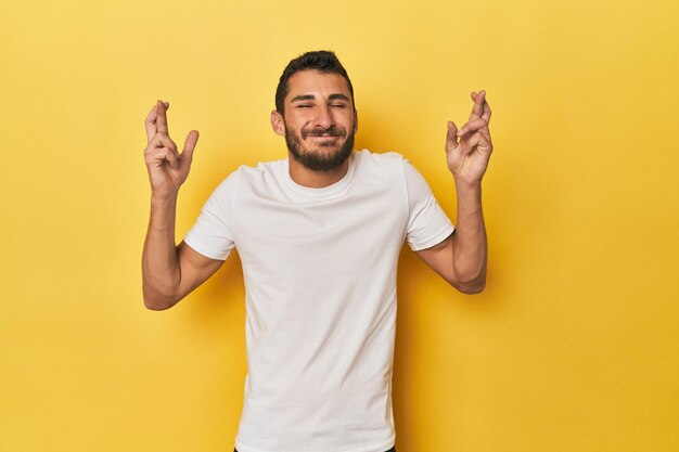 Young Hispanic man on yellow background crossing fingers for having luck