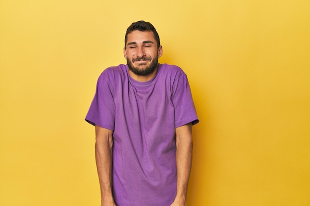 Young Hispanic man on yellow background laughs and closes eyes feels relaxed and happy