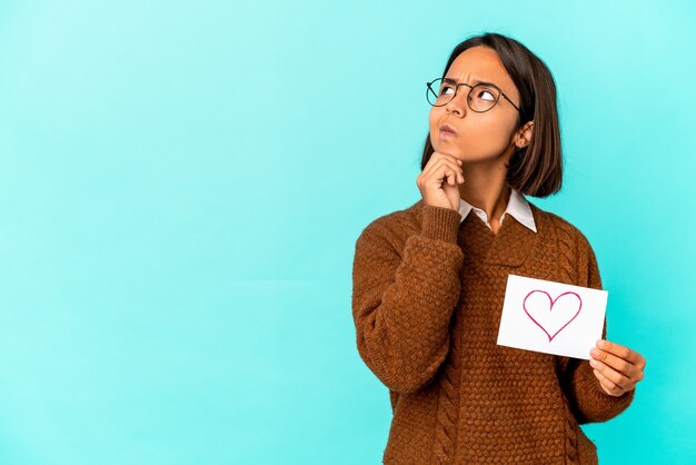 Young hispanic mixed race woman holding a heart paper looking sideways with doubtful and skeptical expression.