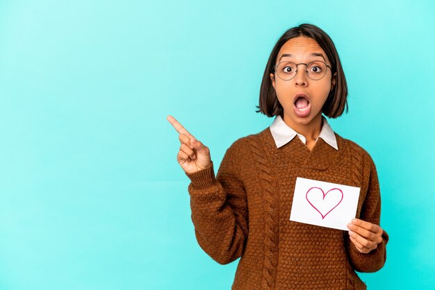 Giovane donna ispanica della corsa mista che tiene una carta del cuore che indica al lato