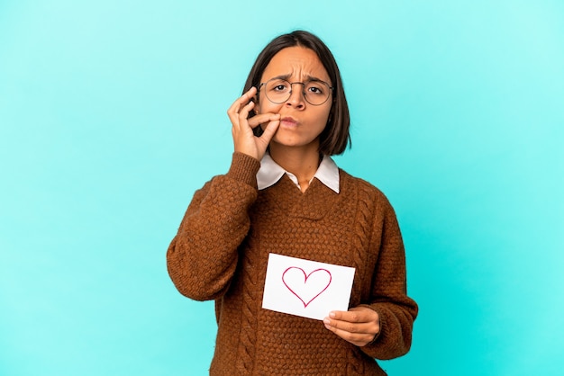 Young hispanic mixed race woman holding a heart paper with fingers on lips keeping a secret.