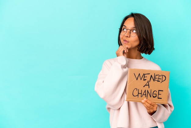 Young hispanic mixed race woman holding an inspiring change message on cardboard looking sideways with doubtful and skeptical expression.