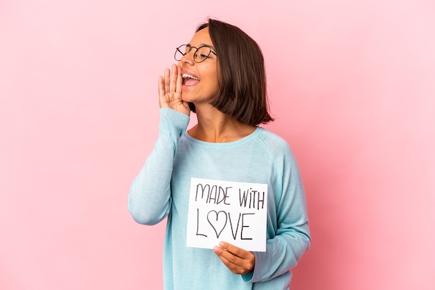 Photo young hispanic mixed race woman holding a made with love paper poster shouting and holding palm near opened mouth.