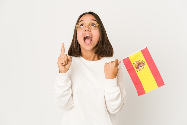 Young hispanic mixed race woman holding a spanish flag pointing upside with opened mouth.