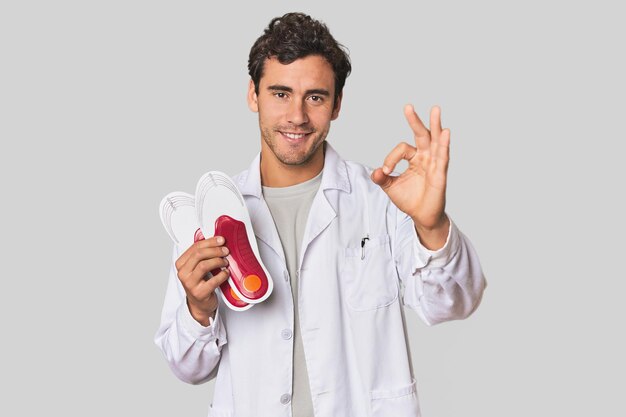 Young Hispanic podiatrist with insoles cheerful and confident showing ok gesture