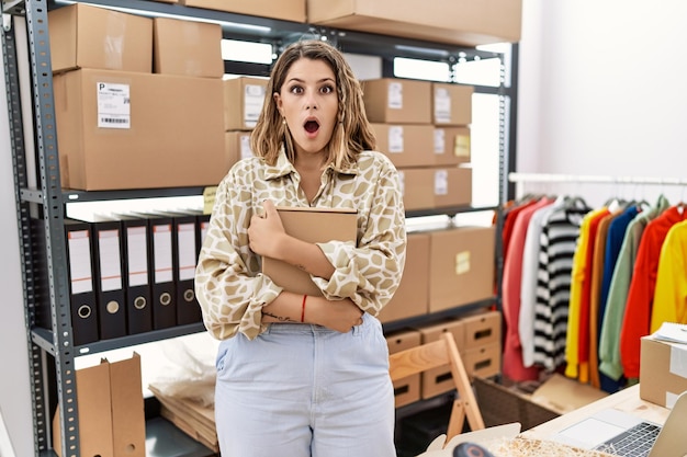 Young hispanic shopkeeper woman holding cardboard box at storeware afraid and shocked with surprise and amazed expression, fear and excited face.