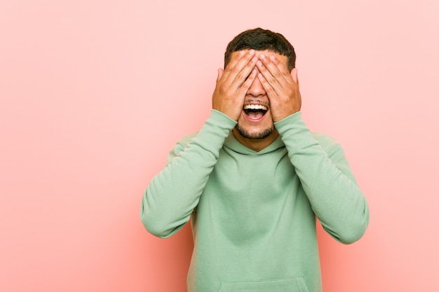 Young hispanic sport man covers eyes with hands, smiles broadly waiting for a surprise.