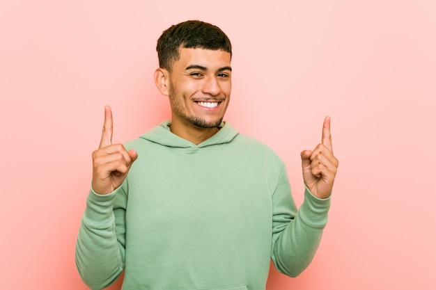 Young hispanic sport man indicates with both fore fingers up showing a blank space.