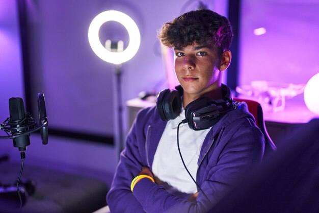 young hispanic teenager streamer smiling confident sitting with arms crossed gesture at gaming room