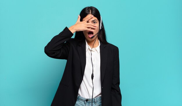 Young hispanic telemarketer woman looking shocked, scared or terrified, covering face with hand and peeking between fingers