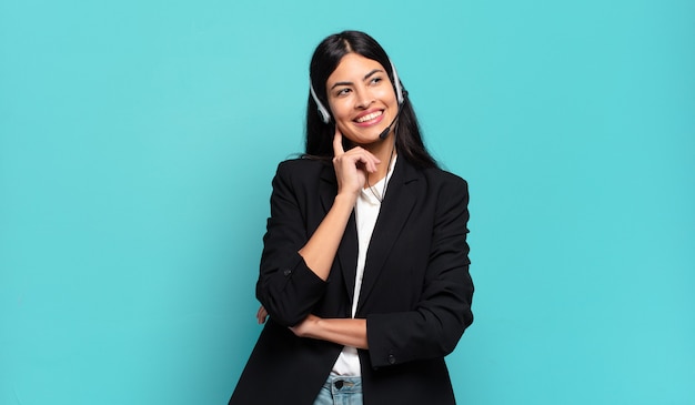 Young hispanic telemarketer woman smiling happily and daydreaming or doubting, looking to the side