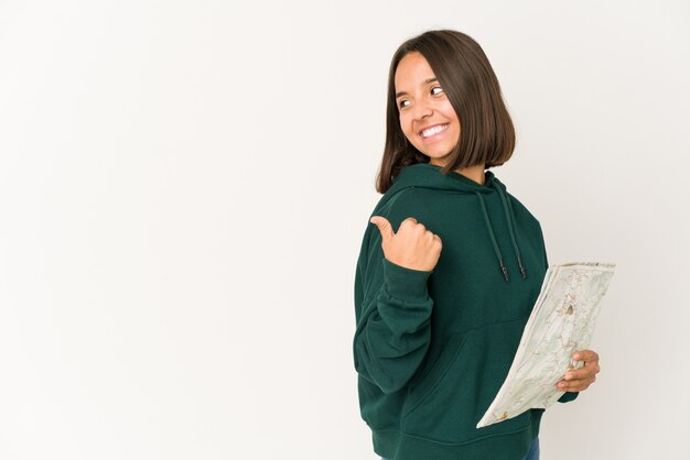 Young hispanic traveler woman holding a map points with thumb finger away, laughing and carefree.