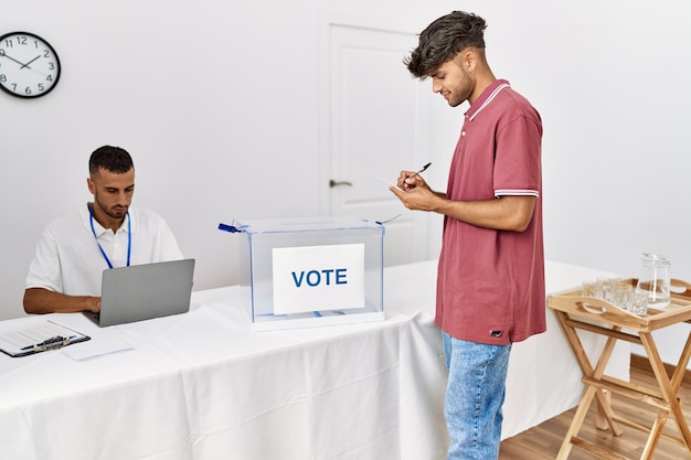 Young hispanic voter man smiling happy writing on vote at electoral center.