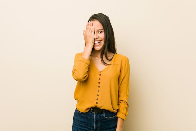 Young hispanic woman against a beige wall having fun covering half of face with palm.