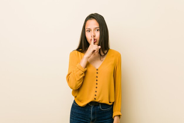 Young hispanic woman against a beige wall keeping a secret or asking for silence.