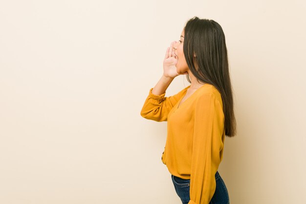 Young hispanic woman against a beige wall shouting and holding palm near opened mouth.