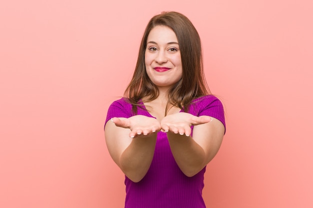 Young hispanic woman against a pink wall holding something with palms