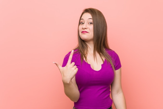 Young hispanic woman against a pink wall pointing with finger at you as if inviting come closer.