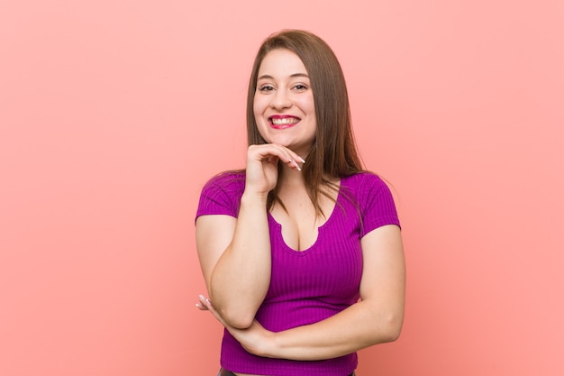 Young hispanic woman against a pink wall smiling happy and confident, touching chin with hand.