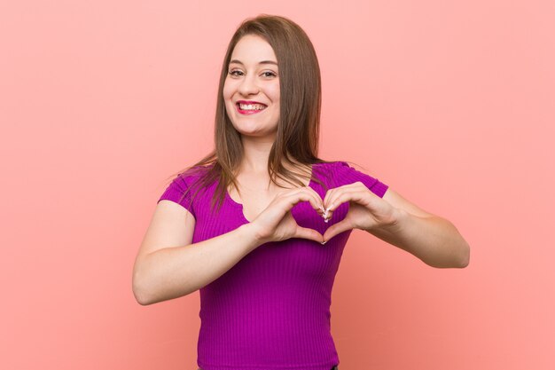 Young hispanic woman against a pink wall smiling and showing a heart shape with hands.