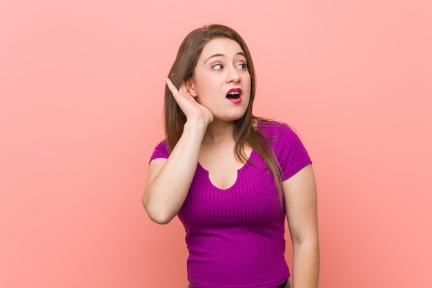 Young hispanic woman against a pink wall trying to listening a gossip.