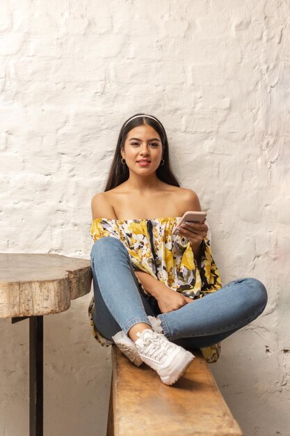 Young hispanic woman casually dressed holding her smart phone and sitting at a rustic wooden table