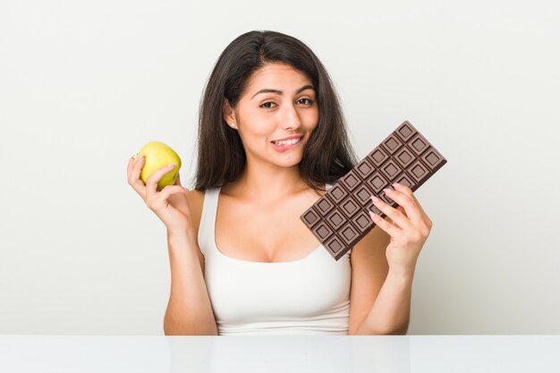 Young hispanic woman choosing between apple or chocolate tablet