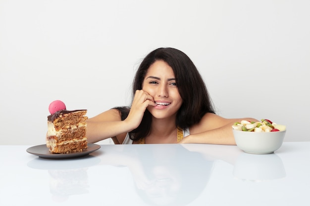 Young hispanic woman choosing between cake or fruit biting fingernails, nervous and very anxious.