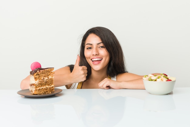 Young hispanic woman choosing between cake or fruit smiling and raising thumb up