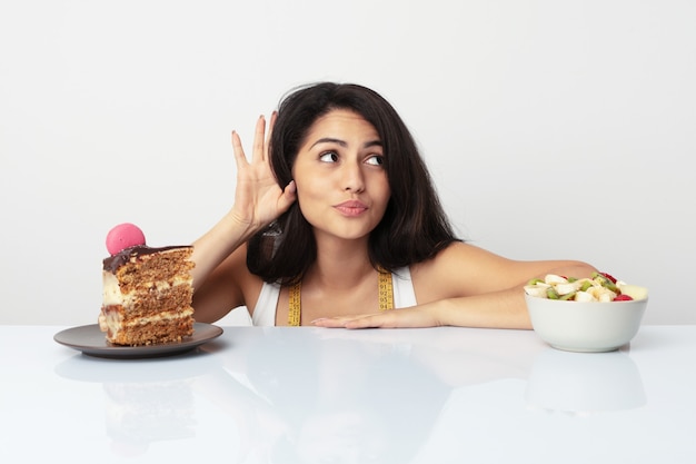 Giovane donna ispanica che sceglie fra la torta o la frutta che prova ad ascoltare un gossip.