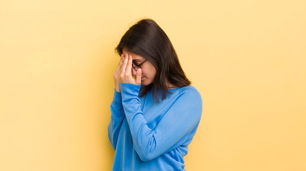 Young hispanic woman covering eyes with hands with a sad frustrated look of despair crying side view