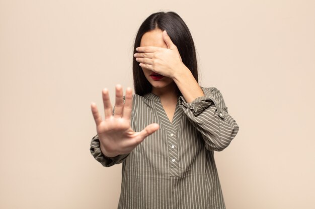 young hispanic woman covering face with hand and putting other hand up front to stop camera, refusing photos or pictures