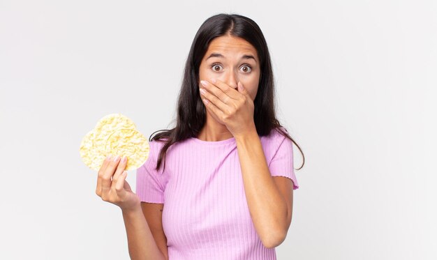 Young hispanic woman covering mouth with hands with a shocked and holding a rice cookie. diet concept