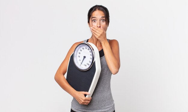 Young hispanic woman covering mouth with hands with a shocked and holding a weight scale. diet concept
