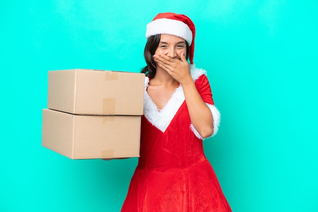 Young hispanic woman dressed as mama noel holding cartoon box\
isolated on blue background happy and smiling covering mouth with\
hand