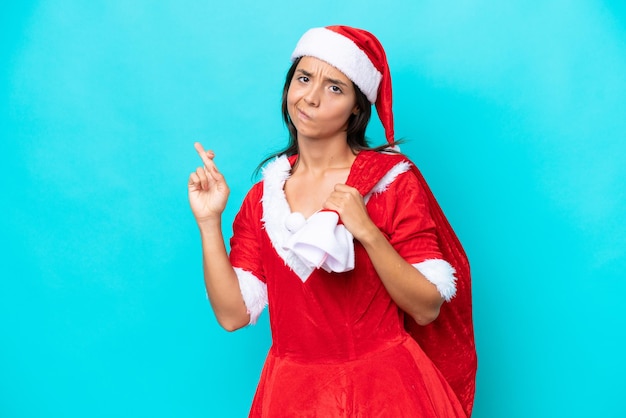 Young hispanic woman dressed as mama noel holding a sack isolated on blue background with fingers crossing and wishing the best