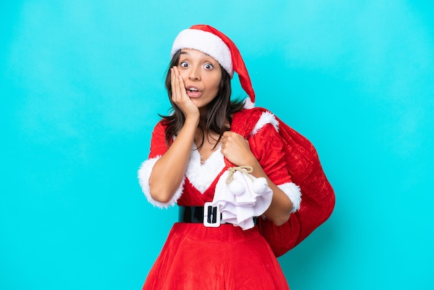 Young hispanic woman dressed as mama noel holding a sack isolated on blue background with surprise and shocked facial expression