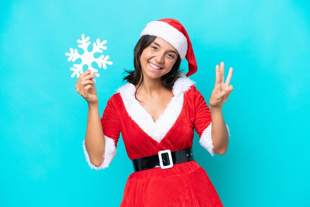 Young hispanic woman dressed as mama noel holding a snowflake isolated on blue background happy and counting three with fingers